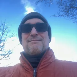 Thad taking a winter selfie outdoors, wearing sunglasses, a beanie, and a red jacket against a blue sky.