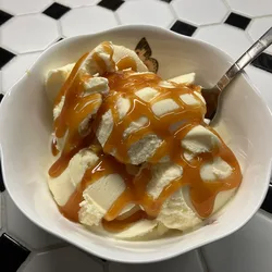 Bowl of vanilla ice cream drizzled with caramel syrup on a black and white tile counter, with a spoon resting in the bowl.