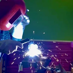 Thad wearing a welding helmet and gloves, sparks lighting up his basement workspace.