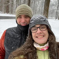 Thad and Jolynn smiling in a snowy wooded area, snowflakes visible on their clothing.