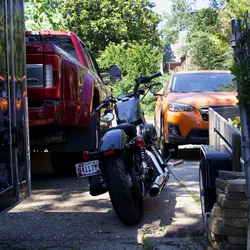 Thad's motorcycle parked between a truck and a trailer, in the shade of the driveway.