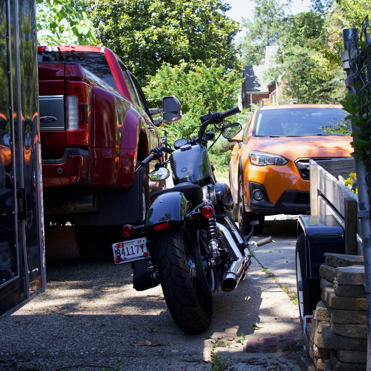 Thad's motorcycle parked between a truck and a trailer, in the shade of the driveway.