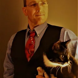 Thad in a vest and tie holding his tuxedo cat, Miss Muffin, in warm indoor lighting.