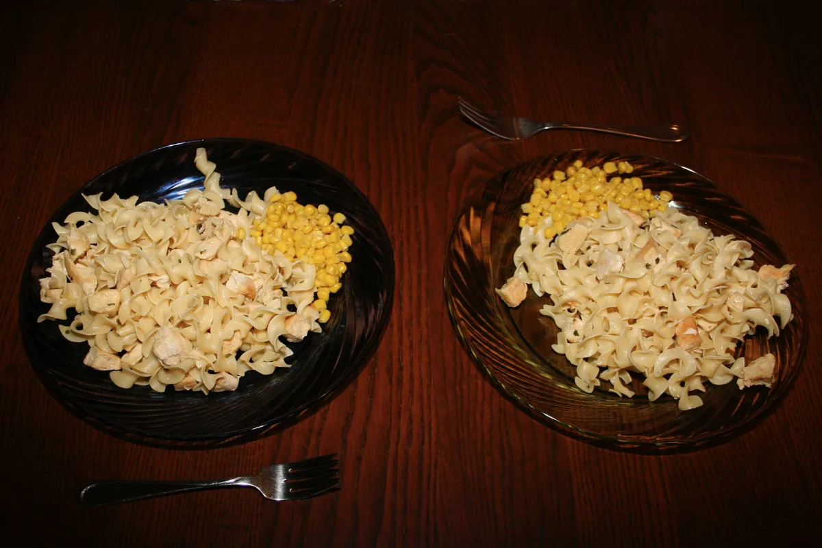 Two plates of chicken and noodles with a side of corn