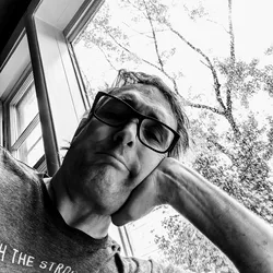 Black and white close-up portrait of me wearing glasses, resting my head on my hand near a rain-covered window with tree branches visible outside.