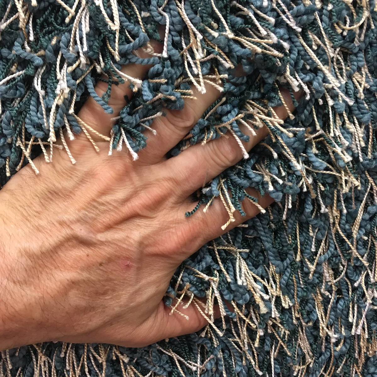 Hand pressing into a long blue-gray shag carpet with thick twisted strands.
