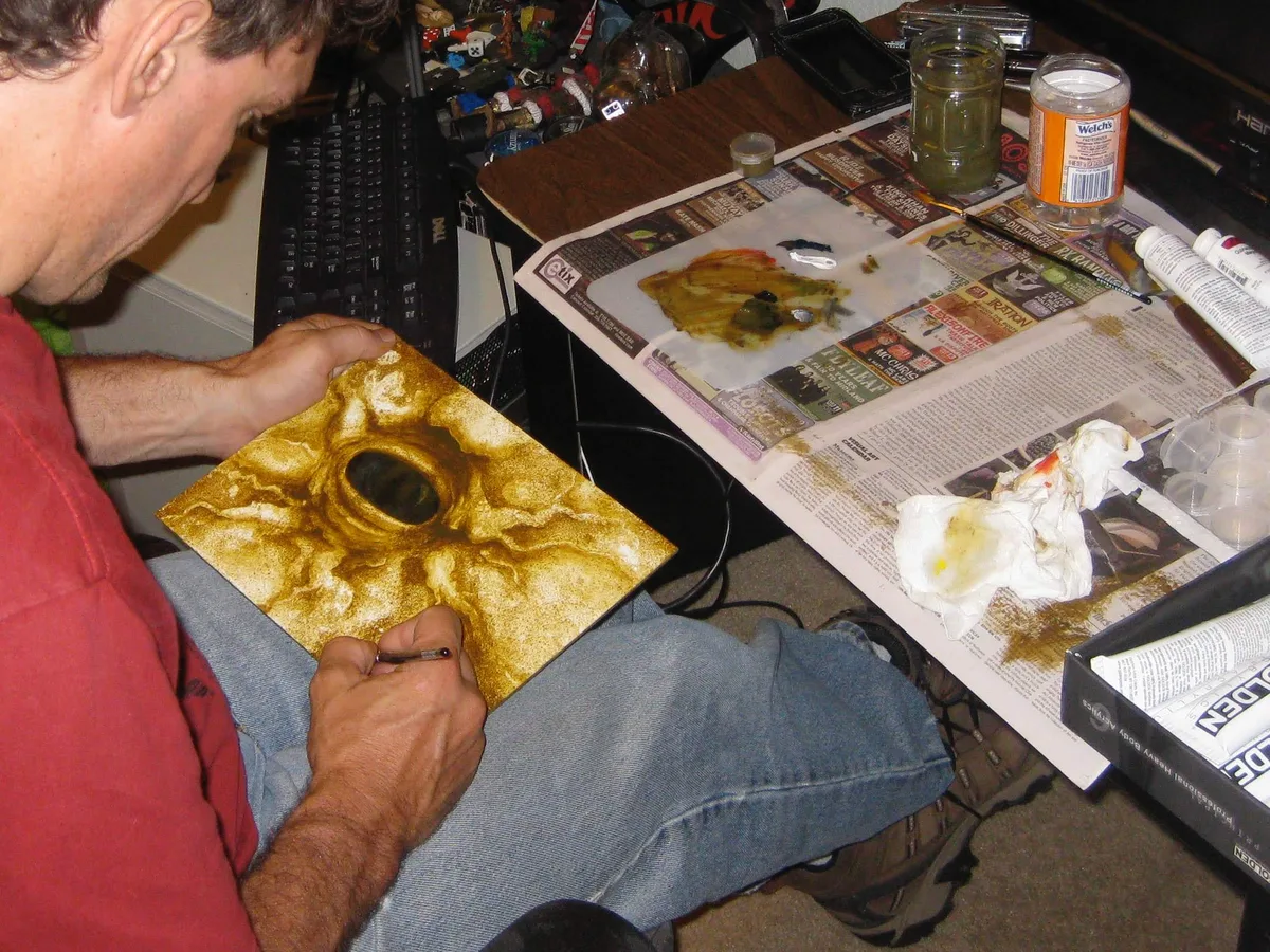 Thad working on the nearly finished Elder's Eye painting on his lap, holding it in one hand while painting with the other.