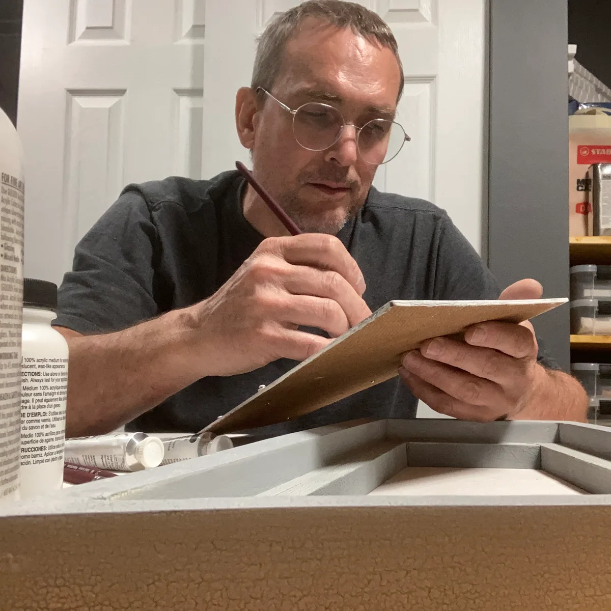 Thad painting on a small wood panel, holding it at an angle with a brush in hand.