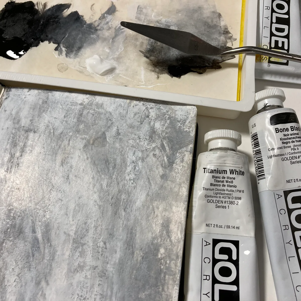 Gray textured underpainting in a sketchbook next to tubes of Titanium White and Bone Black acrylic paint.