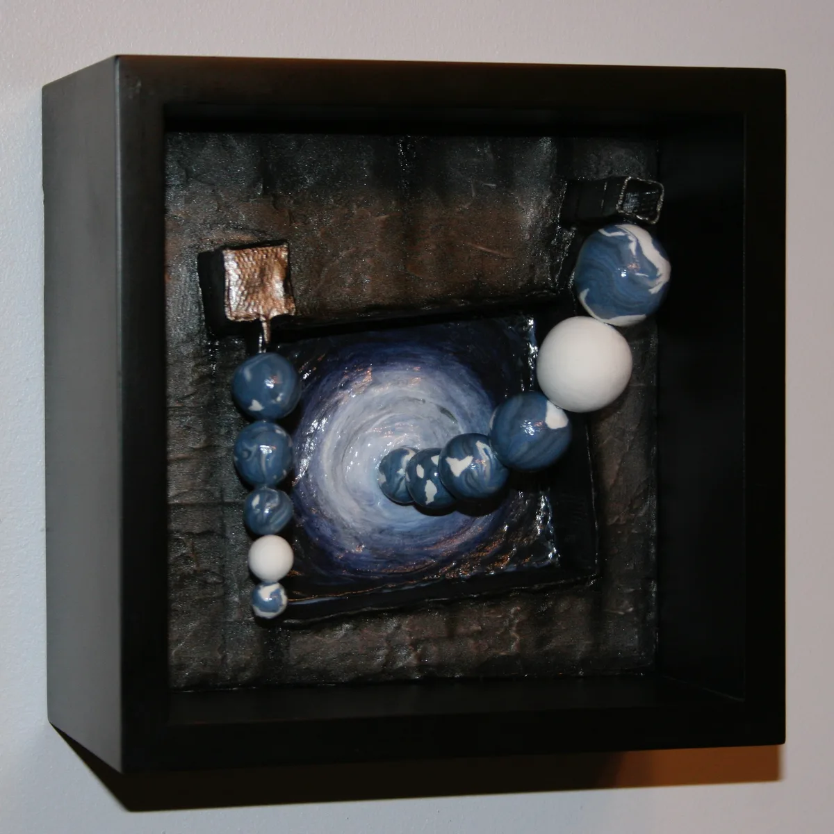 A textured mixed media artwork combining a painted spiral and layered clay beads, mounted in a black shadow box frame.