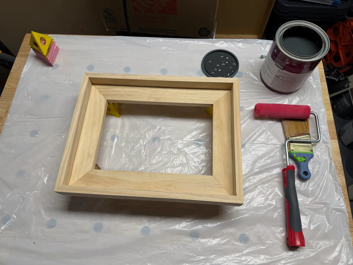 A dried wood frame beside a can of black paint and a foam roller.