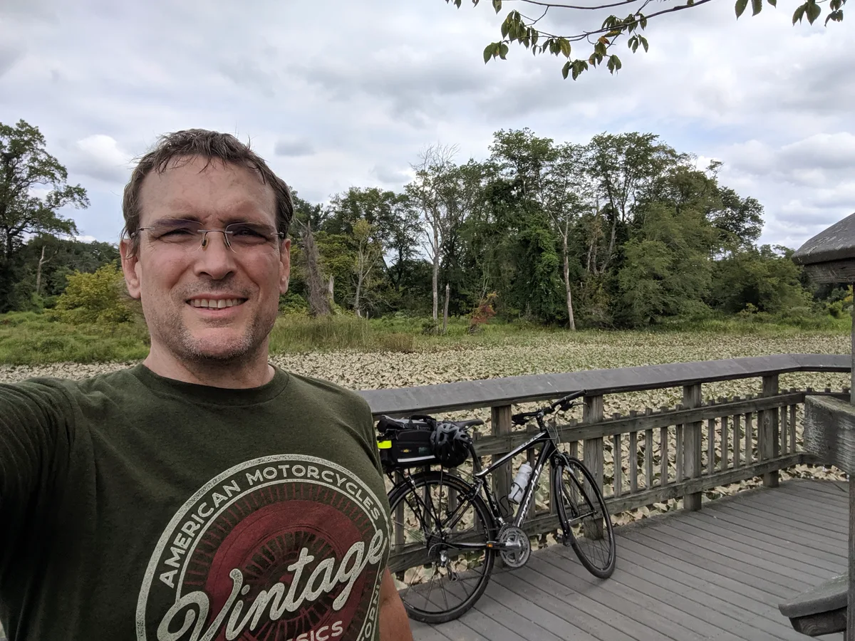 Thad on a boardwalk with his bike, wetland plants filling the water behind him.